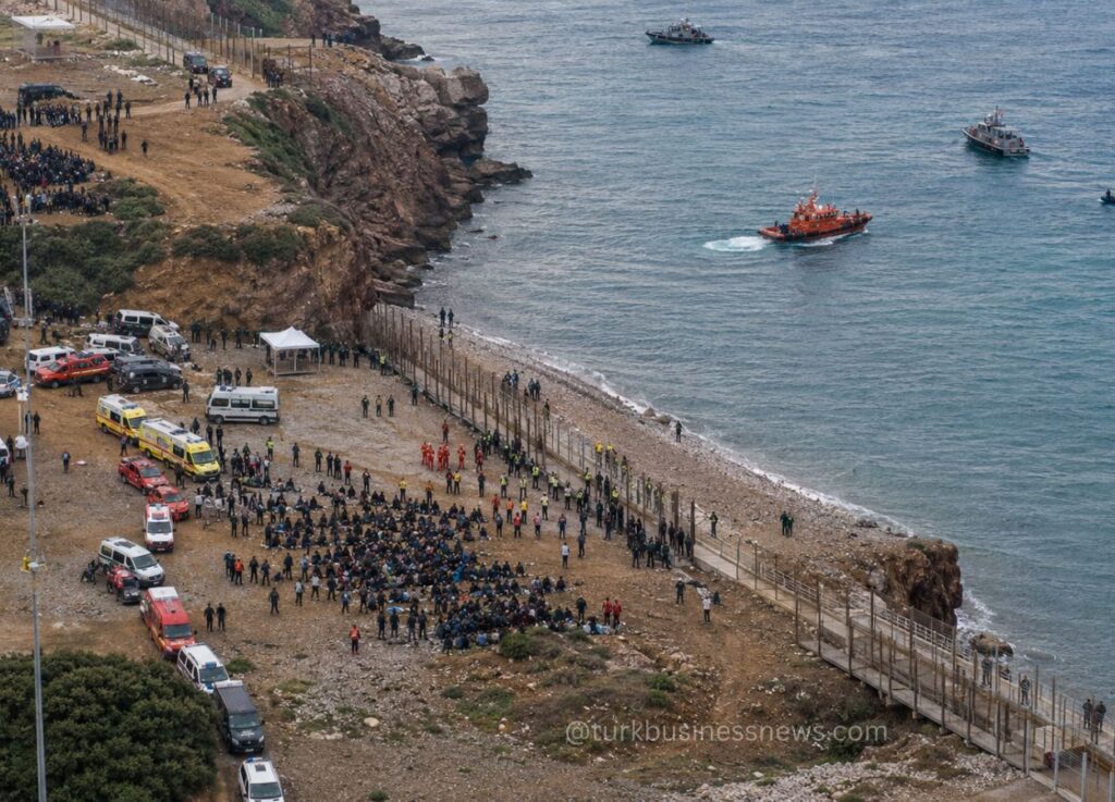 63 - Aerial view of a crowded pebble beach with many people gathered, emergency vehicles parked along the shore, and rescue boats approaching from calm blue sea.