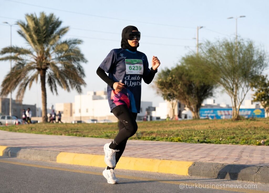 116 - Female runner wearing a hijab and sunglasses mid-stride in a road race, bib 6239, on a sunny street with palm trees in the background.