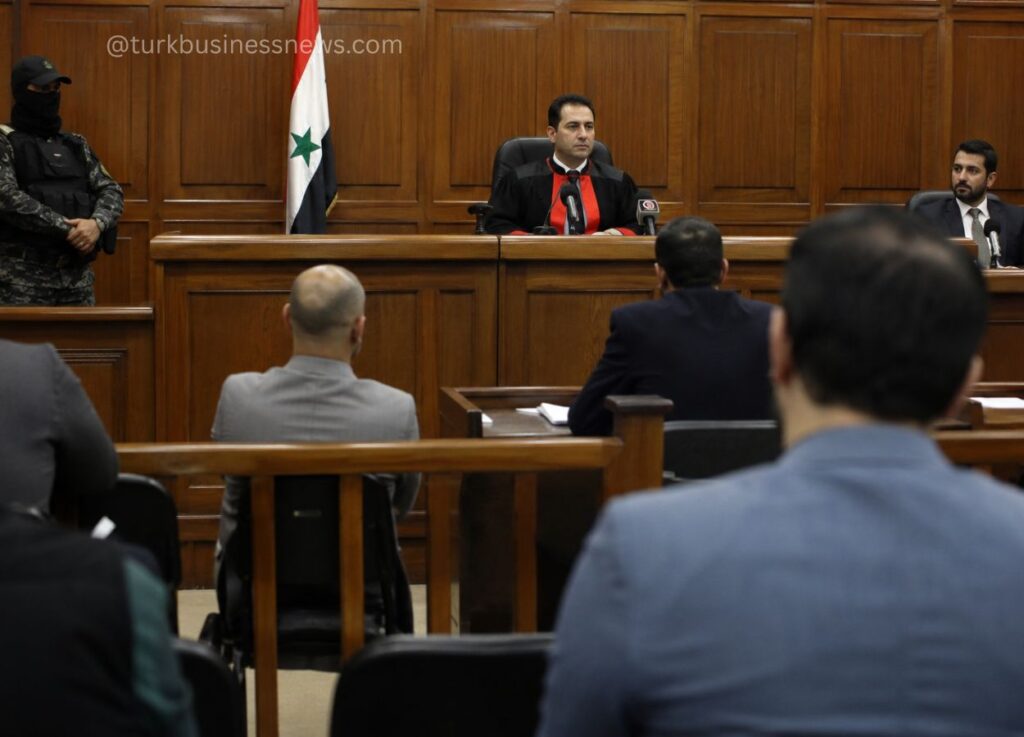 132 - Judge seated at a wooden bench in a courtroom, red-trimmed robe, with a flag behind and attendees facing the panel.