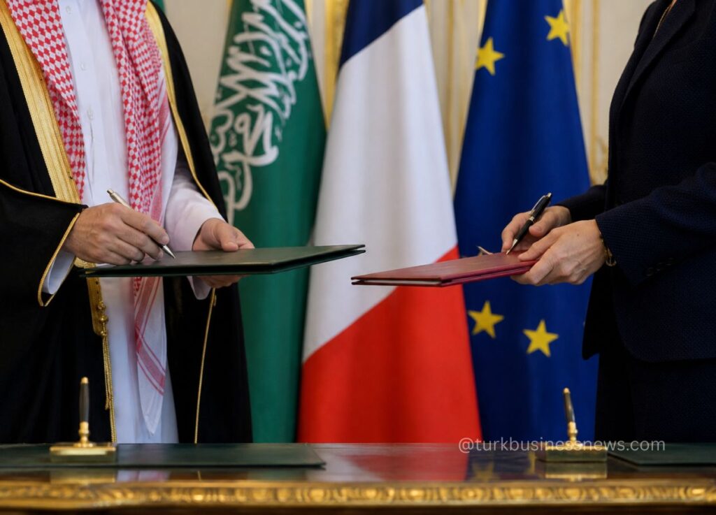 156 - Two officials sign documents on a table as flags of Saudi Arabia, France, and the EU loom behind.