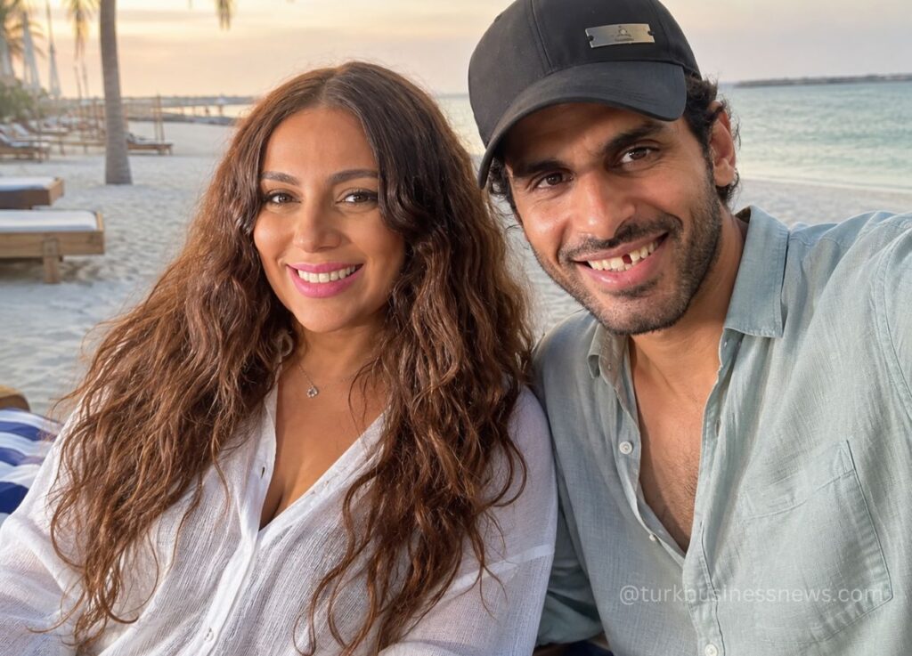 Smiling couple taking a sunset selfie on a sandy beach, with palm trees and the sea in the background.
