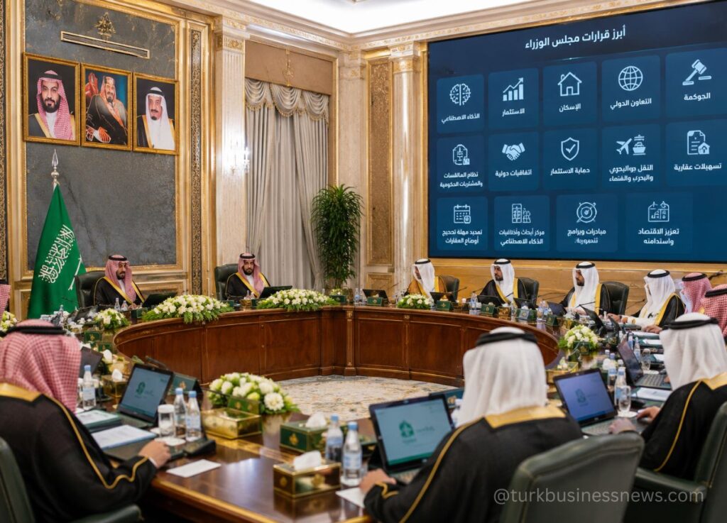 Saudi government ministers sit around a curved wooden conference table in a gilded, ornate room; a Saudi flag and portraits line the wall, with a large blue screen displaying Arabic text behind them.