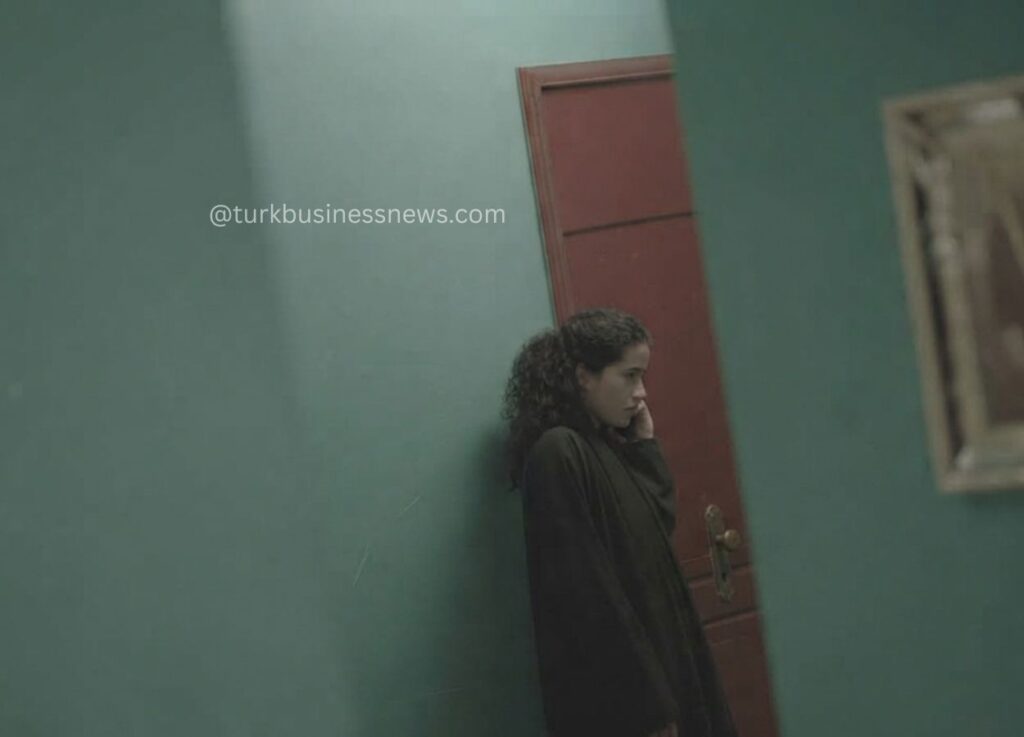 204 - Woman with curly hair in a dark coat leans against a green wall beside a red door, looking pensive.